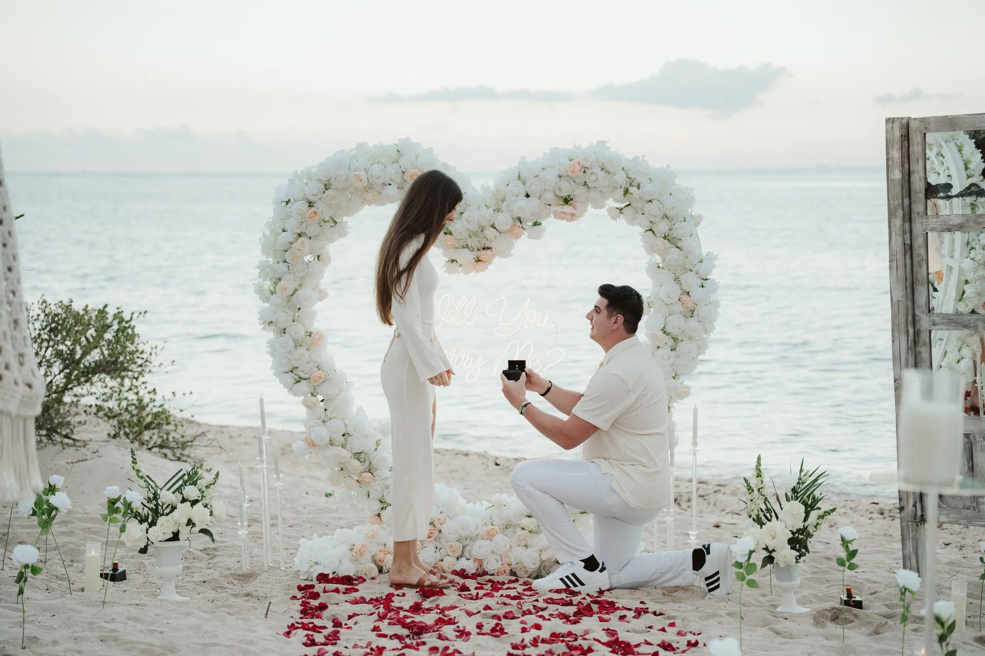 Pareja en el momento de una propuesta al atardecer en playa, rodeada de flores blancas y el Caribe