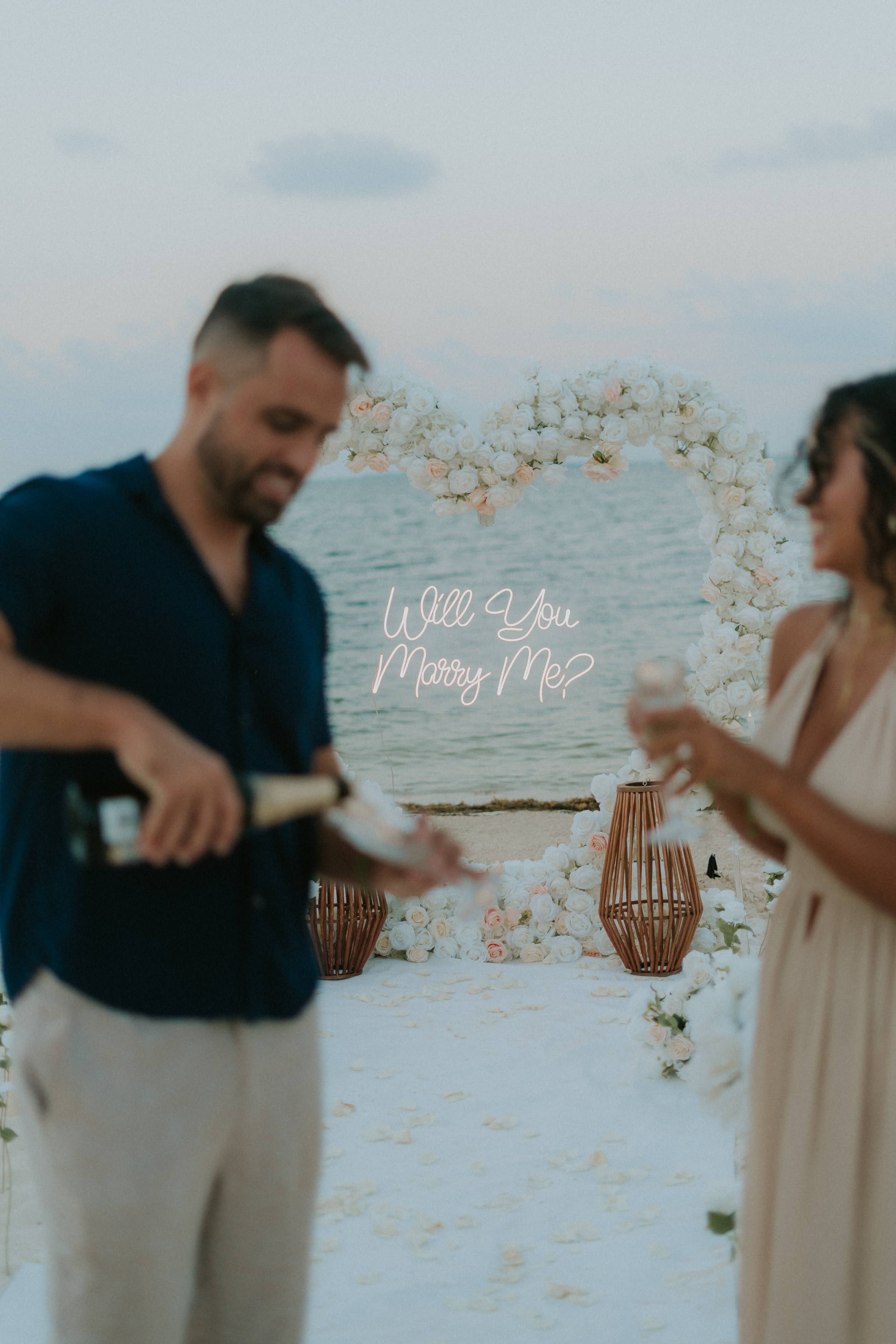 Intimate marriage proposal on a secluded emerald-water beach in Cancun featuring a white floral heart arch and romantic picnic setup on the sand.