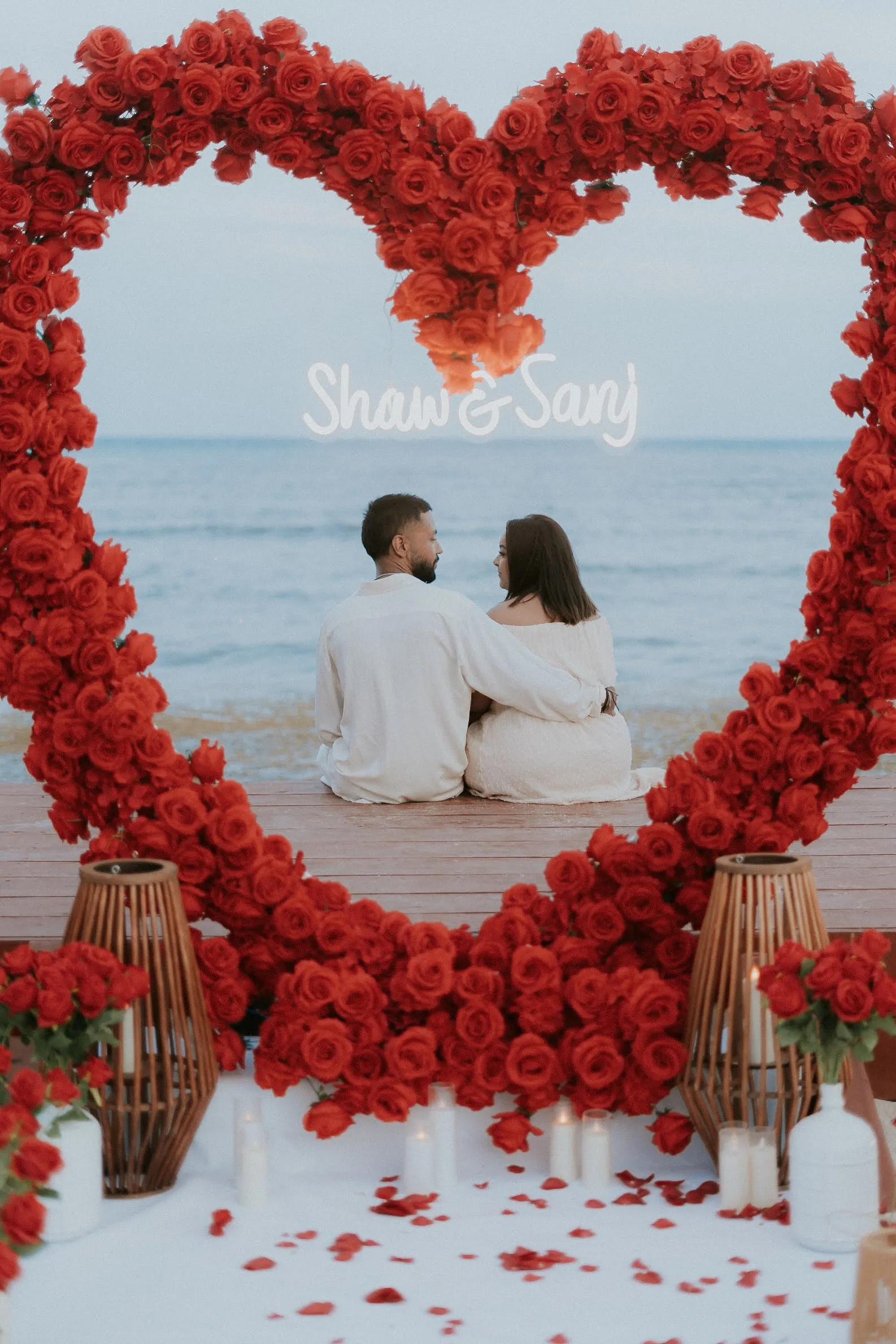 Pareja celebrando su compromiso frente al Caribe, enmarcada por un arco de corazón de rosas rojas
