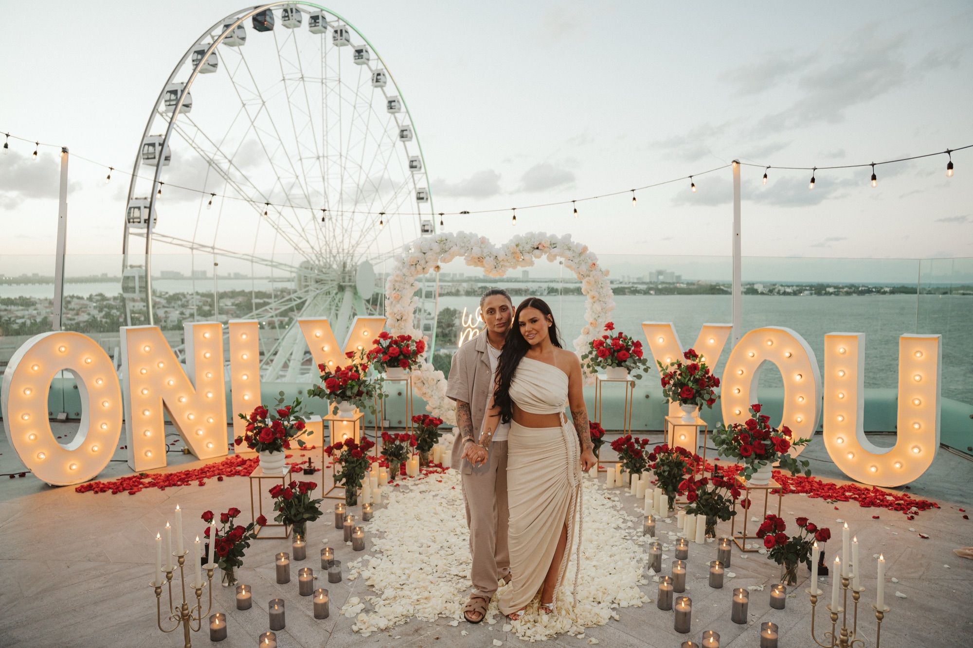 Bespoke rooftop proposal with the couple in front of custom "ONLY YOU" marquee letters, floral heart arch, and ocean view