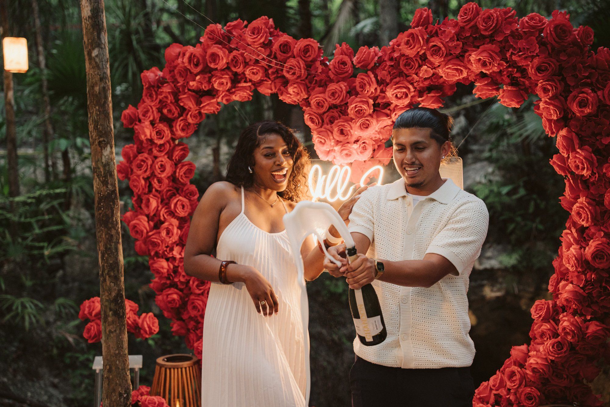 Joyful couple popping champagne in a cenote after a successful surprise proposal, celebrating their engagement in a tropical paradise.