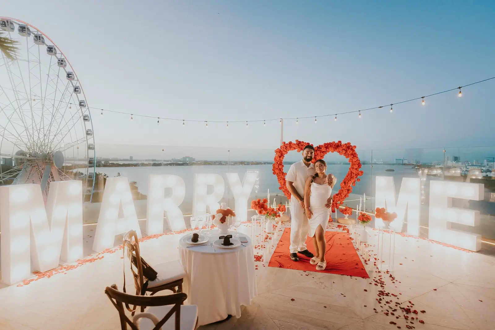 Rooftop marriage proposal celebration with flowers and illuminated marry me letters