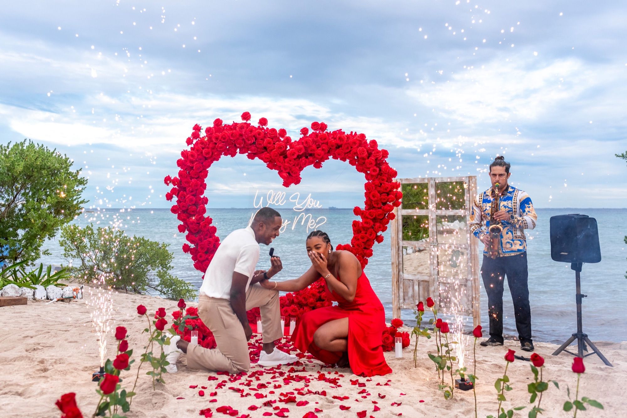 Signature beach proposal moment with red rose heart arch, live saxophone, and cold sparklers